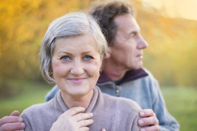 older woman smiling with husband behind her looking off into distance